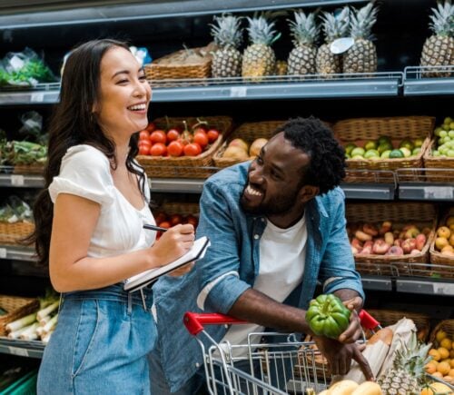 A couple laughing in the fruit and vegetable aisle of a supermarket, pushing a shopping cart