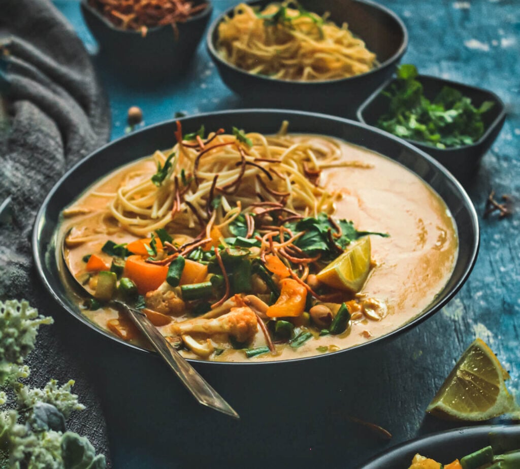 A black ceramic bowl filled with a vegan khow suey soup recipe and topped with crunchy noodles and other garnishes